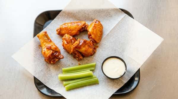 Four glazed chicken wings, celery sticks, and a small cup of white dipping sauce on parchment paper.