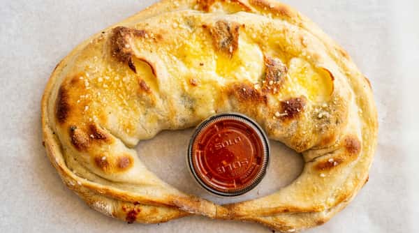 Calzone with golden crust, melted cheese, and a side of marinara sauce in a plastic cup.