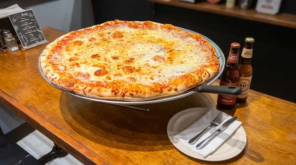 Large cheese pizza on a serving stand with two beer bottles and a plate with a napkin on a wooden table.