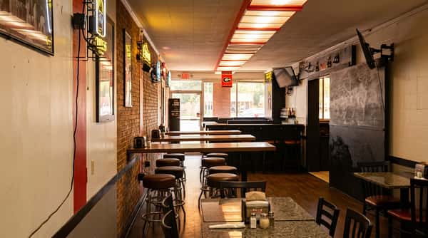 A narrow, dimly lit bar or restaurant interior with booth seating and tables, bright light streaming from the far end.