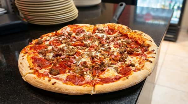 Meat lover's pizza with pepperoni, sausage, and ham on a black countertop, next to stacked plates.
