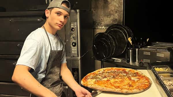 Chef holding a large pizza with mozzarella, pepperoni, and sausage in a commercial kitchen.