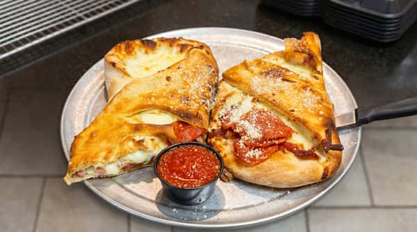 Two calzones, one with pepperoni and the other with ham and salami, served with a side of marinara sauce on a metal pizza pan.