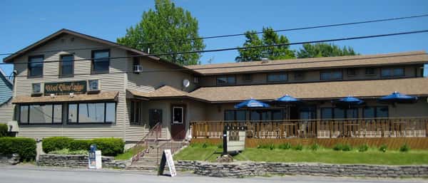 Exterior view of West View Lodge, a rustic American restaurant and lodge featuring a spacious front patio with blue umbrellas, set against a clear blue sky and surrounded by greenery.