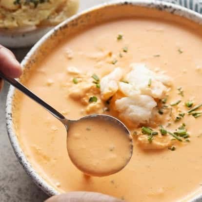 "Creamy seafood bisque served in a rustic bowl, topped with tender chunks of lobster or shrimp and fresh herbs, with a spoon dipping into the rich orange soup.
