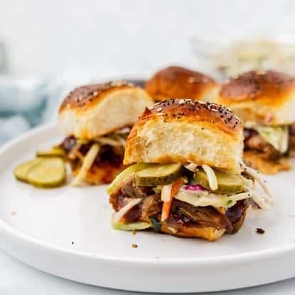 BBQ pulled pork sliders topped with coleslaw and pickles on soft, toasted buns, served on a white plate—ideal for casual dining, party platters, or American-style comfort food menus.
