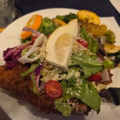 Crispy breaded cutlet topped with a fresh garden salad of mixed greens, cherry tomatoes, red onion, and shredded parmesan, garnished with a lemon wedge—vibrant and hearty entrée.