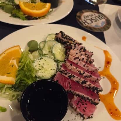 Seared sesame-crusted ahi tuna slices served with cucumber salad, green olives, orange wedge, and soy dipping sauce—vibrant seafood appetizer with Asian fusion flair.