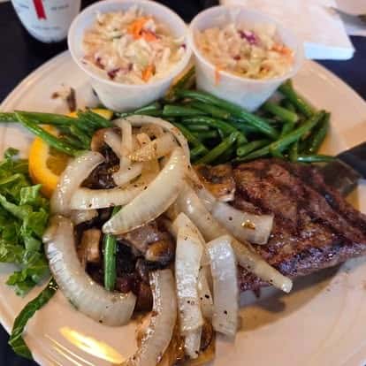 Flame-grilled steak topped with sautéed onions and mushrooms, served with green beans, crisp lettuce, orange slice, and two sides of creamy coleslaw—classic American steakhouse plate.