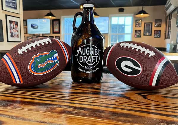 A growler from Auggie's Draft Room is flanked by two college footballs—one for the Florida Gators and the other for the Georgia Bulldogs—setting the scene for a spirited game day rivalry at this Saint Augustine spot.