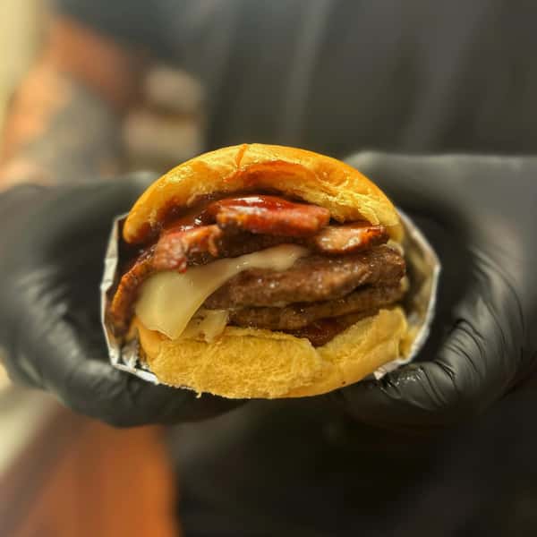 Juicy double cheeseburger with thick bacon slices and barbecue sauce held by gloved hands.