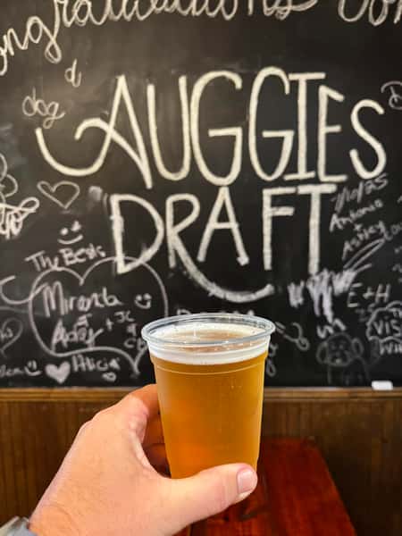 Hand holding a plastic cup of draft beer in front of a chalkboard wall with "Auggie's Draft" and doodles.