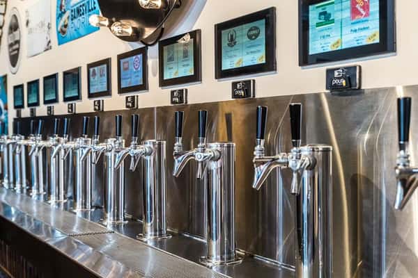 Close-up of chrome beer taps beneath digital screens at Auggie's Draft Room self-pour wall.