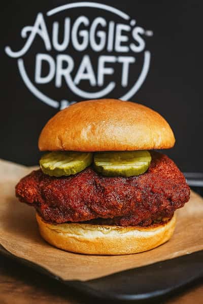 Spicy fried chicken sandwich topped with pickles on a toasted bun in front of an Auggie's Draft Room sign.