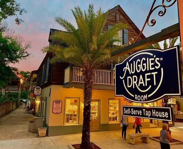 Auggie's Draft Room sign glows at sunset outside a self-serve tap house surrounded by palm trees and cozy storefronts.