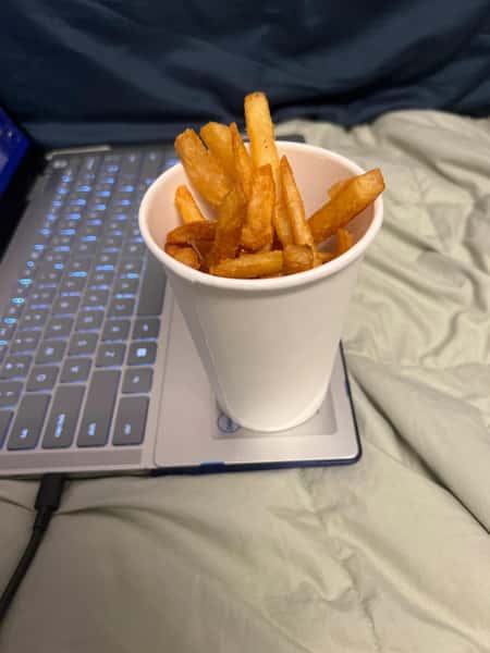 Golden French fries in a white paper cup placed on a laptop keyboard over a light-colored blanket.