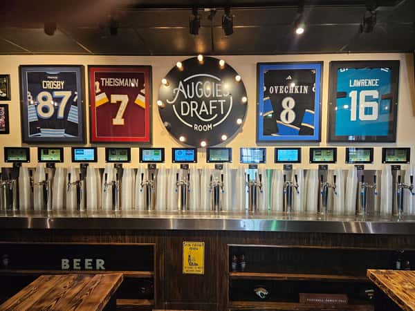 Beer tap wall at Auggie's Draft Room with self-pour screens, plastic cups, and framed sports jerseys above the taps.