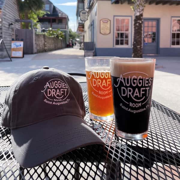 Two branded Auggie's Draft Room pints—one amber, one dark—sit on an outdoor table alongside a matching gray baseball cap, with a quaint St. Augustine street and palm tree in the background.