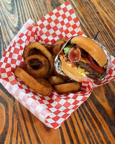 This mouthwatering basket features a classic bacon cheeseburger stacked with lettuce, tomato, and melted cheese—paired perfectly with a golden helping of crispy onion rings, all served on a red-and-white checkered liner atop a rustic wooden table.