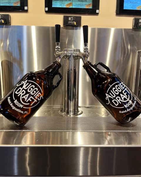 A pair of branded Auggie's Draft Room growlers are propped on a sleek stainless steel beer tap station, ready to be filled with fresh draft brews.
