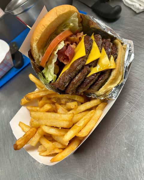 Cheeseburger stacked with four beef patties, bacon, lettuce, tomato, and fries in a tray on a stainless steel counter.