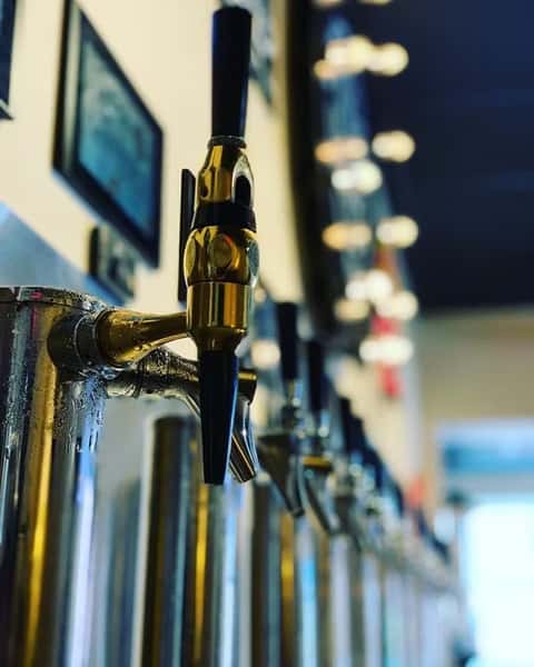 Close-up of a gold beer tap handle at a self-pour draft wall, with soft lights and other taps blurred in the background.