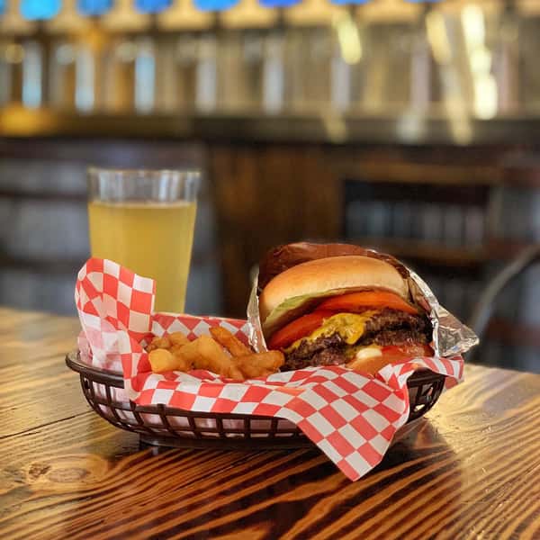 Cheeseburger with tomato, lettuce, and pickles served with fries in a basket alongside a pint of light beer on a wooden table.