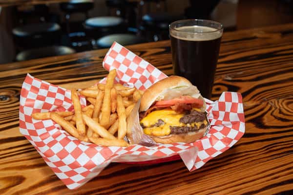 Cheeseburger with lettuce, tomato, and fries served in a red basket with a pint of dark beer on a woodgrain table.