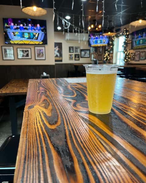 A cold light beer in a plastic cup on a rustic woodgrain table, with sports on TV screens and holiday lights decorating the cozy pub interior.
