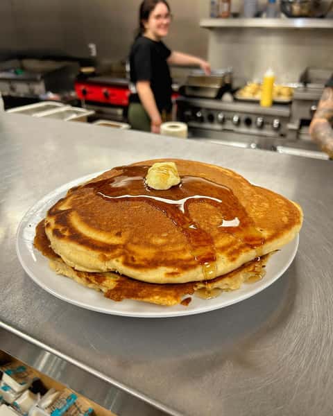 Stack of fluffy pancakes topped with a pat of butter and maple syrup, served on a white plate in a diner kitchen.