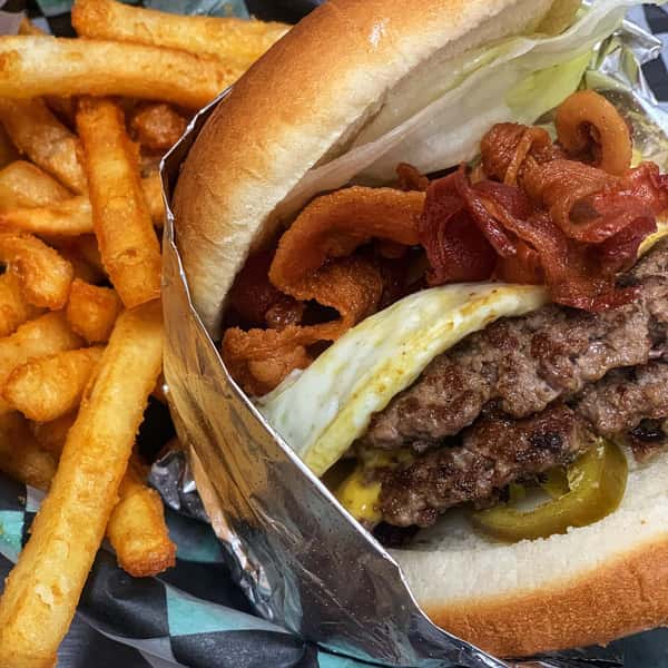 Close-up of a bacon cheeseburger with fried egg, jalapeños, and lettuce, served with crispy golden fries.