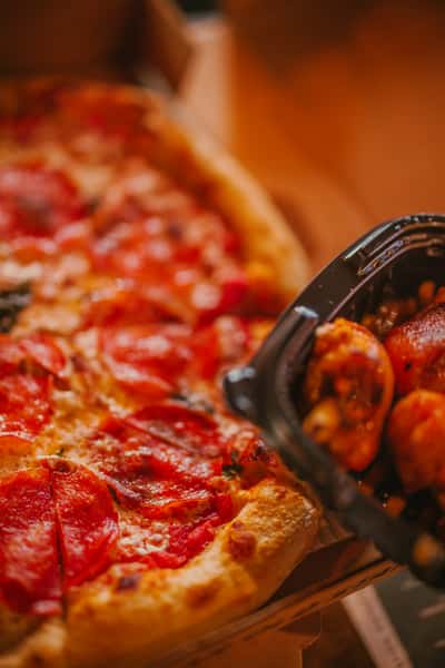 Close-up of a pepperoni pizza with a golden crust next to a takeout container filled with saucy chicken wings, emphasizing classic pizzeria comfort food.