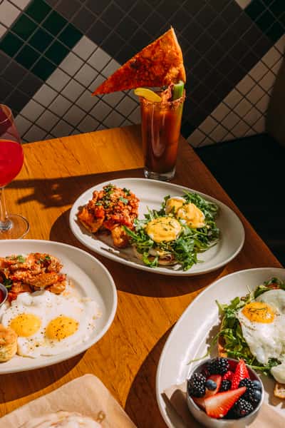 Colorful brunch spread featuring eggs Benedict over arugula, sunny-side-up eggs with crispy potatoes, fried chicken, fresh berries, and craft cocktails including a Bloody Mary garnished with a slice of pizza.