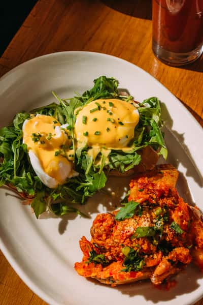 Poached eggs with hollandaise sauce on a bed of arugula served with a side of roasted potatoes topped with a savory tomato-based sauce.
