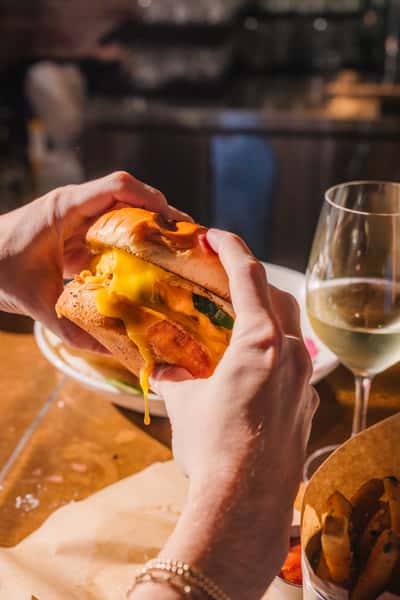 Hands holding a juicy cheeseburger with melted cheddar oozing between toasted buns, paired with crispy fries and a glass of white wine at a trendy gastropub.