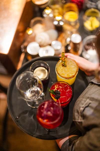 Cocktail tray with colorful craft drinks including a yellow pineapple margarita with chili rim, a berry garnish red cocktail, and a purple spritz, served in a lively bar setting