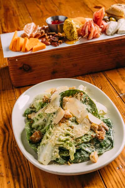 Fresh Caesar salad with crisp romaine hearts, shaved Parmesan, garlic croutons, and creamy dressing served alongside a gourmet charcuterie board with cured meats, cheeses, nuts, and fig jam on a rustic wooden table.