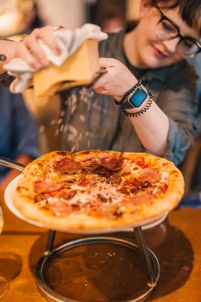 Close-up of a freshly baked meat-lovers pizza topped with prosciutto, sausage, and mushrooms, as a server grates parmesan cheese over it at a cozy Italian restaurant.