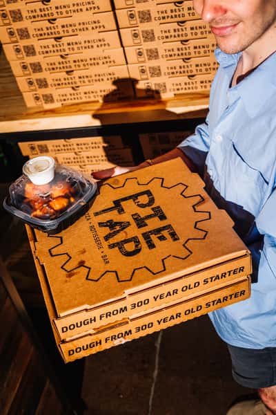Pizza takeout from Pie Tap Pizzeria, featuring branded boxes highlighting 'dough from 300-year-old starter' alongside a container of saucy wings with ranch, ready for delivery or pickup.