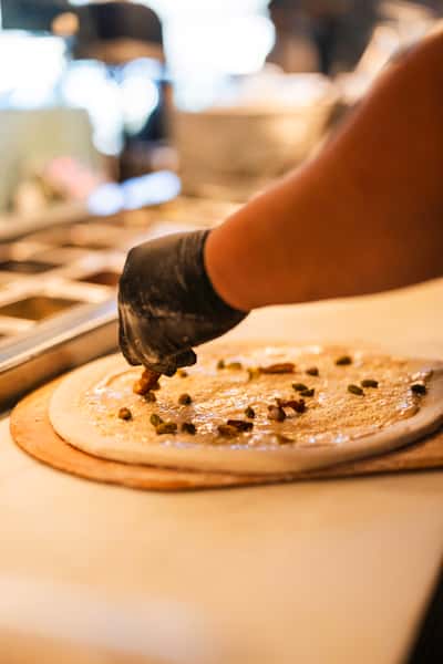 Close-up of a gloved chef hand-topping fresh pizza dough with sliced jalapeños and seasoning in an open kitchen, showcasing artisanal pizza-making in progress.
