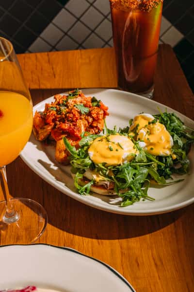 Brunch plate with eggs Benedict topped with hollandaise and arugula alongside spicy breakfast potatoes, with a mimosa and Bloody Mary in the background.