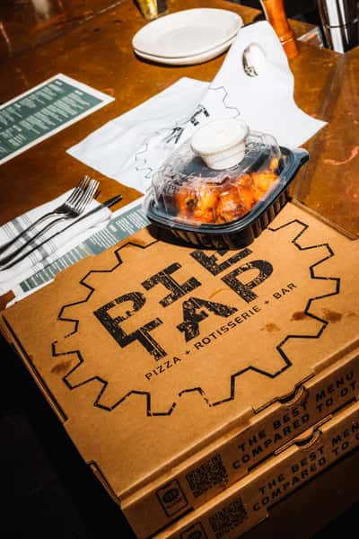 Branded Pie Tap pizza boxes with a sealed takeout container of saucy buffalo wings and ranch dressing, placed on a rustic bar table with utensils and plates nearby.