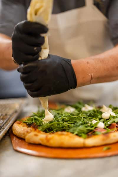 Freshly baked arugula pizza topped with dollops of creamy white sauce, being finished by a chef wearing black gloves in a gourmet kitchen.