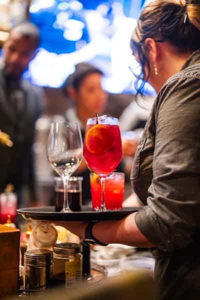 Server carrying a tray of colorful cocktails, including a red spritz with orange slice and stemware, at a bustling bar with blurred patrons in the background.