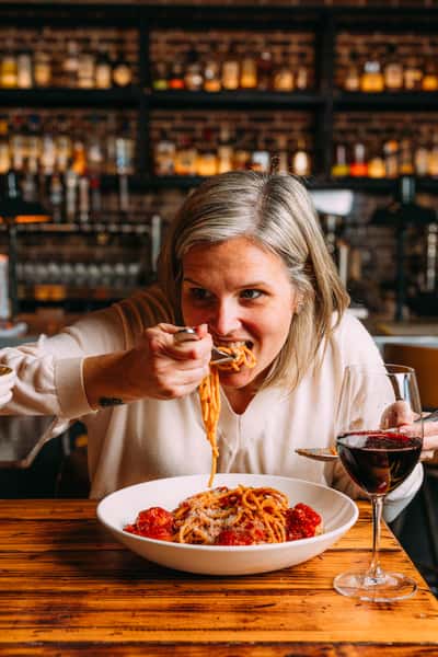Woman enjoying a bowl of spaghetti and meatballs topped with Parmesan cheese, paired with a glass of red wine in a warm, upscale Italian restaurant.