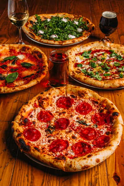 Four gourmet pizzas with various toppings including pepperoni, arugula, and basil, served with wine and beer on a rustic wooden table.