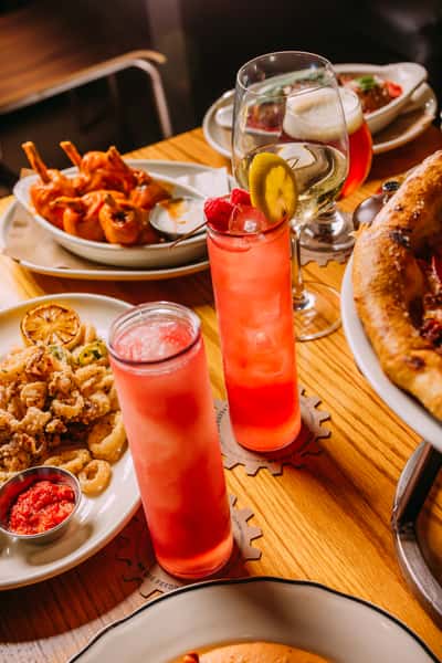 Tabletop spread featuring raspberry cocktails with lemon and cherry garnish, glasses of wine and craft beer, paired with crispy fried calamari, saucy buffalo wings, and a freshly baked pizza at a lively American eatery.