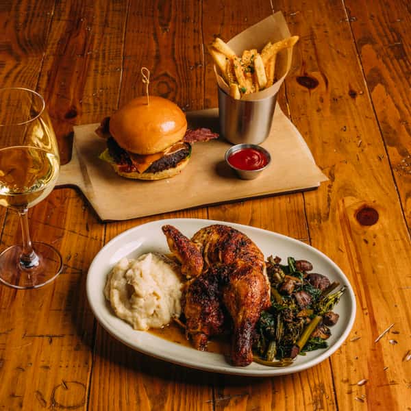 Gourmet meal with roasted chicken, mashed potatoes, sautéed greens, and a bacon cheeseburger with truffle fries served with white wine.