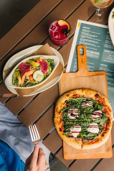 Wood-fired arugula and burrata pizza with a drizzle of balsamic glaze served on a wooden paddle alongside a fresh garden salad and a vibrant berry cocktail on a patio table.