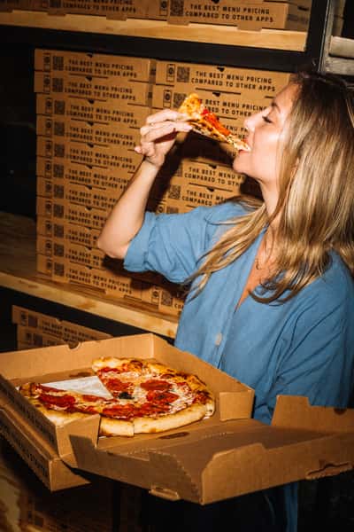 A woman in a blue shirt enjoys a slice of pepperoni pizza from a cardboard box, standing in front of a stack of branded pizza boxes at a casual pizzeria.